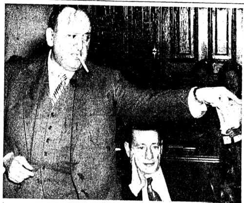 Newspaper photograph of a man in a suit and tie standing and pointing to his left. Another man is seated next to him.